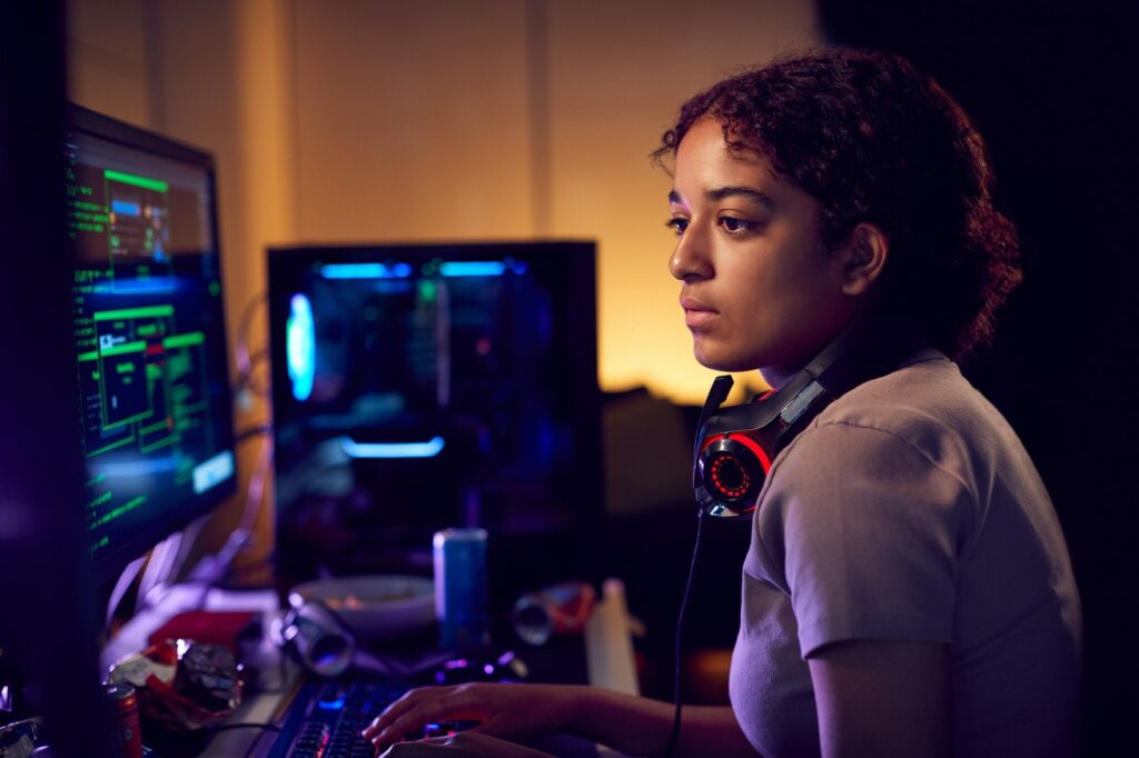 Female Teenage Hacker Sitting In Front Of Computer Screens Bypassing Cyber Security