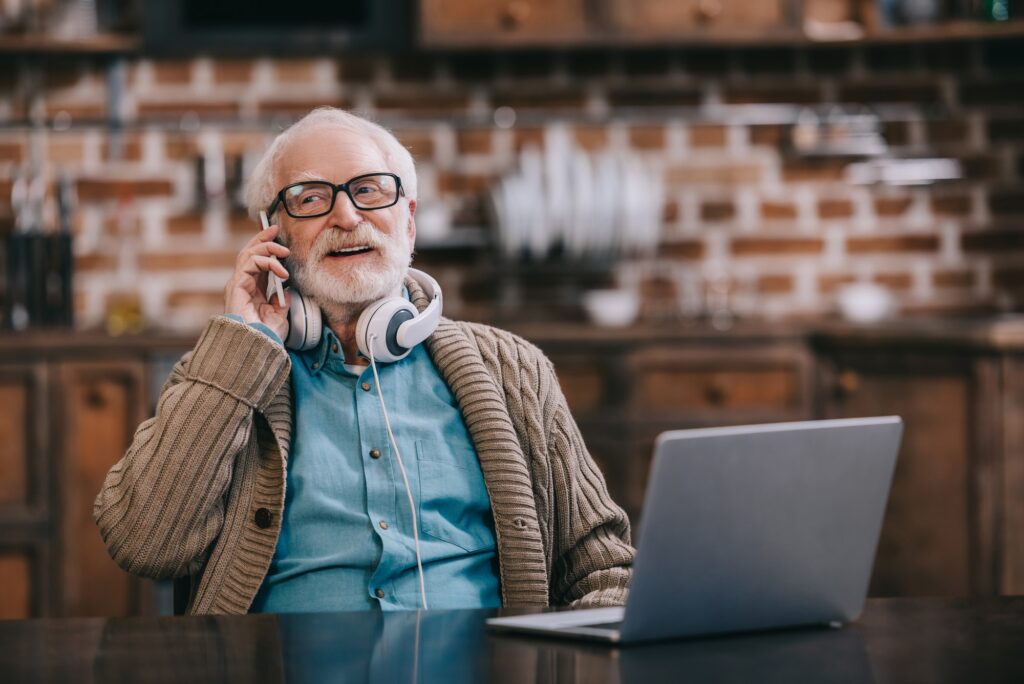 Old man in headphones talking on phone by laptop